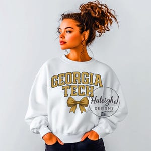 May include: White sweatshirt with gold "GEORGIA TECH" text and a gold bow graphic. A circular logo with "Haleigh J DESIGNS & Co." is also visible. A casual, comfortable top.