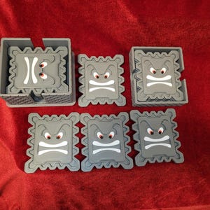 Op de afbeelding: Set van zes grijze onderzetters met een cartoon-ontwerp met een boos gezicht. De onderzetters zijn gestapeld in een toren met een grijze basis.