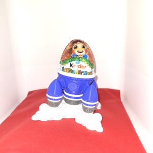 May include: A blue and white rocket-shaped holder with a Kinder Surprise egg inside. The egg features a cartoon animal on the front and the text "Kinder Surprise".