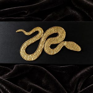 Peut inclure: Une boîte rectangulaire noire ornée d'un emblème de serpent en métal doré texturé, centré sur le couvercle. Le serpent est représenté dans une position enroulée en forme de S avec une finition de surface détaillée et rugueuse.