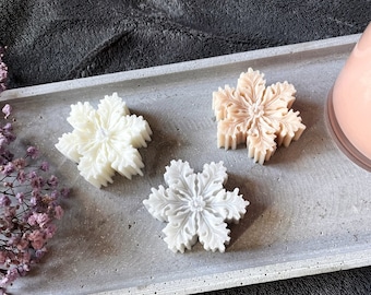 Puede incluir: Tres velas decorativas con forma de copo de nieve en una bandeja gris. Las velas son blancas, marrón claro y marrón oscuro.