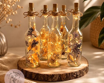 May include: Five clear glass bottles with cork stoppers, each containing dried flowers and warm white fairy lights. Each bottle is tied with a brown twine bow at the neck. The bottles are arranged on a round wooden slice. A logo with the text Deko-Kennerin is visible.