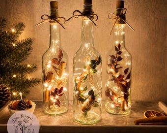 Op de afbeelding: Drie doorzichtige glazen flessen met kurken, elk omwikkeld met touw en gevuld met warmwitte kerstverlichting, kaneelstokjes en gedroogde bladeren. De flessen zijn van binnenuit verlicht en zorgen voor een zachte gloed.