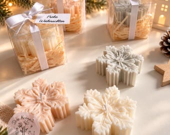 May include: Three snowflake-shaped candles in shades of white and light peach. Two candles are packaged in clear square boxes with white ribbons and tags reading Frohe Weihnachten, while one white and one peach candle sit unwrapped.
