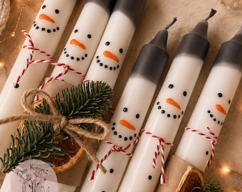 May include: A set of six white taper candles featuring hand-painted snowman faces with orange carrot noses and black dot eyes and mouths. Each candle has a black top, a red and white twine tie, and is wrapped in brown paper with a dried orange slice.