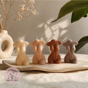 May include: Four torso-shaped candles in varying shades of beige, tan, brown, and taupe are arranged in a row on a textured, off-white ceramic tray. Each candle features a single white wick at the top and depicts a sculpted human upper body.