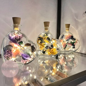 Könnte beinhalten: Drei herzförmige Glasflaschen mit Korkstopfen, gefüllt mit Trockenblumen und Lichterketten. Die Flaschen sind in einer Reihe auf einer reflektierenden Oberfläche angeordnet.
