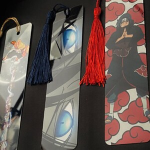 May include: Three different bookmarks with colorful tassels. The first bookmark features a blue and white eye design. The second bookmark features a red and white cloud design with a black figure in the center. The third bookmark features a yellow and white design with a figure in the center.