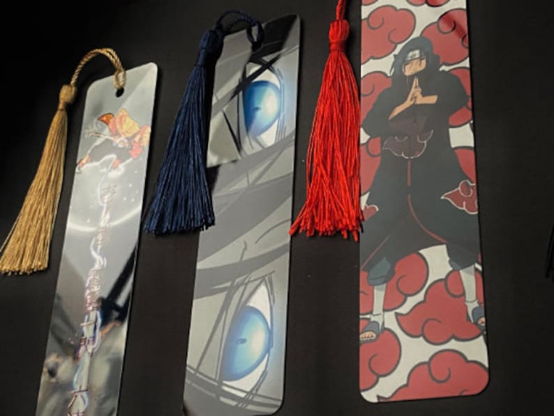 May include: Three different bookmarks with colorful tassels. The first bookmark features a blue and white eye design. The second bookmark features a red and white cloud design with a black figure in the center. The third bookmark features a yellow and white design with a figure in the center.