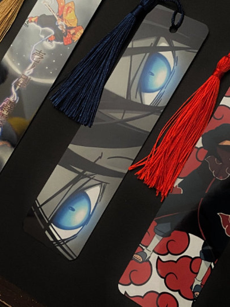 May include: Three different anime bookmarks with colorful tassels. The first bookmark features a character with a red and yellow outfit. The second bookmark features a close-up of a character's eye with a blue iris. The third bookmark features a character with red clouds and a black and red outfit.