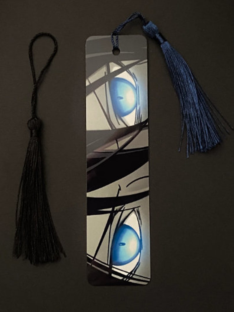 May include: A black and white bookmark with a close-up of a blue eye. The bookmark has a black tassel and a blue tassel.