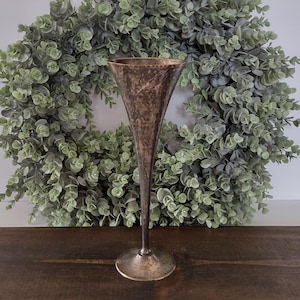 May include: A tall, slender, metallic goblet with a flared, wide rim and a narrow stem leading to a circular base. The surface of the vessel features a dark, mottled, and textured finish with visible signs of oxidation and aged patina.
