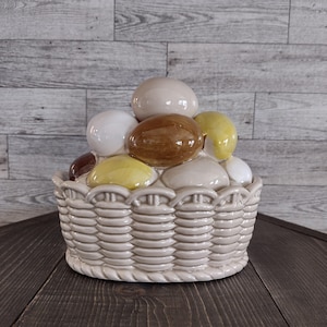 May include: A ceramic basket filled with decorative eggs. The basket is a light beige color with a woven texture. The eggs are various colors, including white, brown, and yellow. The basket is sitting on a dark wooden surface.