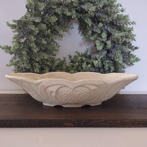 May include: An elongated, cream-colored ceramic bowl with a scalloped rim and a textured, embossed pattern on the exterior. The bowl features a glossy finish and a symmetrical, oval shape, resting on a flat base with a slightly raised center.