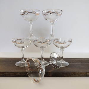 May include: A set of six clear glass coupe glasses with stems and circular bases. Each glass features a delicate gold-colored wheat or grain pattern around the rim and a thin gold band along the edge of the bowl, arranged in a tiered display.