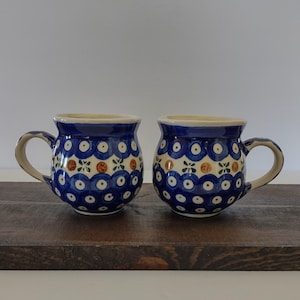 May include: Two ceramic mugs with a blue and white floral pattern. The mugs have a rounded shape with a handle and a cream-colored rim. The design includes blue circles, brown accents, and floral motifs. They sit on a dark wooden surface.