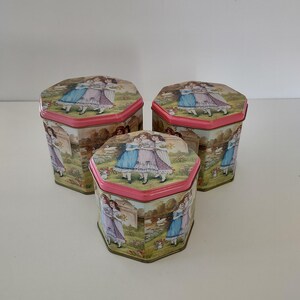 May include: Three hexagonal metal tins with pink lids and bases. Each tin features a printed illustration of three figures in blue, pink, and white dresses standing in a garden setting with swans and greenery. The tins are arranged in a triangular formation.