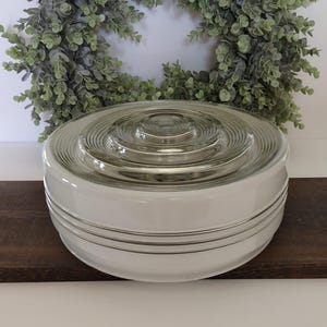 May include: A round, white glass light fixture cover featuring a clear, concentric circular pattern on the top surface. The sides of the fixture are smooth and opaque white, accented by two thin, horizontal silver-toned bands encircling the base.