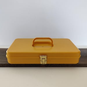 May include: A rectangular mustard yellow plastic storage case featuring a centered integrated handle and a single metallic gold-toned latch mechanism on the front edge. The container has smooth, rounded corners and a flat top surface design.