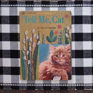 May include: A vintage children's book titled "Tell Me, Cat" by Ellen Fisher. The cover features a detailed illustration of a fluffy orange cat and embroidered floral elements. The book rests on a black and white checkered tablecloth.