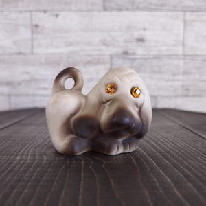 May include: A ceramic dog figurine in a seated position, featuring a cream and brown color scheme. The dog has large, amber-colored rhinestone eyes and a curled tail with a hole. The figurine is on a dark wooden surface.