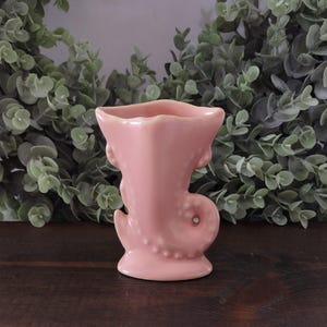 May include: A small, pink ceramic vase with a unique, curved design. The vase has a flared top and a base with a decorative, tentacle-like shape. The vase is approximately 5 inches tall.