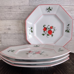 May include: White octagonal plates with a red trim and a strawberry design. The plates have a central image of strawberries and floral accents. A stack of three plates is in the foreground, with another plate visible in the background.