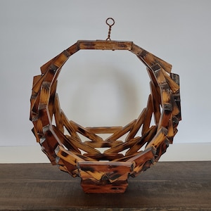 May include: A circular, collapsible wooden basket featuring a lattice design with a scorched, dark brown finish. The structure is composed of interconnected wooden slats and includes a small metal loop at the top for hanging purposes.