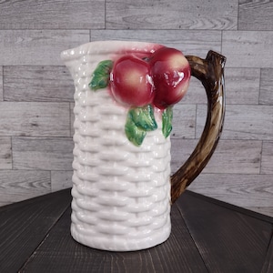May include: White ceramic pitcher with a basket weave design. The pitcher features a brown handle and is adorned with a cluster of red and yellow apples and green leaves. The pitcher is a decorative item.