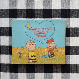 May include: A vintage children's book titled "You're In LOVE, Charlie Brown" featuring illustrations of Charlie Brown and other Peanuts characters. The cover has a blue background with a heart-shaped speech bubble. The book is set against a black and white checkered background.