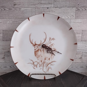 May include: A decorative plate repurposed as a clock, featuring a brown deer illustration. The white plate has brown accents and a decorative clock mechanism. The clock is displayed on a metal stand, adding a rustic touch to the design.