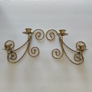 May include: A pair of gold-toned metal wall sconces, each featuring two candle holders supported by curved, swirling wire arms. The design includes circular scroll accents at the ends of the metal framework, creating a symmetrical appearance.