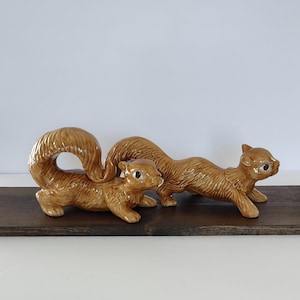 May include: Two ceramic squirrel figurines in a warm brown glaze. One squirrel has a large, curved tail, while the other is in a running pose. Both have small, dark eyes and are positioned on a dark wooden surface.
