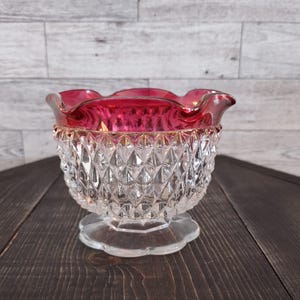 May include: A clear glass bowl featuring a diamond-cut pattern on the exterior and a ruffled, deep red rim. The bowl sits on a circular, scalloped base, showcasing a combination of transparent glass and a vibrant red upper edge with a wavy design.