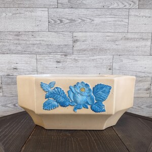 May include: A cream-colored hexagonal ceramic planter featuring a raised, hand-painted blue rose design on the front panel. The rose has a small yellow center and is surrounded by detailed blue leaves, providing a textured, three-dimensional appearance.
