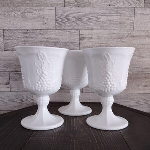 May include: Three white milk glass goblets featuring a raised, embossed pattern of grape clusters and leaves on the bowl. Each goblet has a sturdy pedestal base with a textured, beaded stem design and a smooth, flared rim for drinking.