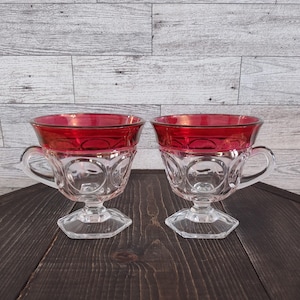 May include: Two clear glass cups with a deep red band around the top rim. Each cup features a circular embossed pattern on the body, a sturdy handle on the side, and a hexagonal base. The glass is transparent with a smooth, polished finish.