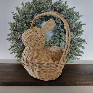 May include: A light tan woven wicker basket shaped like a rabbit with a tall arched handle. The basket features a round body and a single upright ear, with a small pink circular accent on the side of the head and subtle pastel green and pink woven detailing around the base.