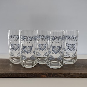May include: A set of five clear glass tumblers, each featuring a blue speckled heart pattern and a matching speckled band near the rim. The glasses have a cylindrical shape with a slightly raised base, showcasing a consistent decorative motif.