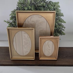May include: A set of three rectangular gold-toned picture frames, each featuring an oval-shaped inner matting. The frames are arranged in a tiered formation, with the largest in the back and two smaller frames positioned in the front.