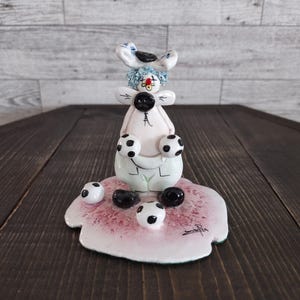 May include: A whimsical ceramic figurine of a clown holding soccer balls. The clown has a white body, blue hair, and a red nose. The figurine sits on a pink base with additional soccer balls scattered around it. The base has a signature.
