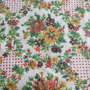 May include: A white fabric featuring a repeating cross-stitch pattern of floral clusters in shades of mustard yellow, dark brown, and bright red, accented by vibrant green leaves and stems, with small red cross-stitch dots filling the negative space.