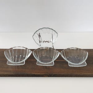 May include: Four clear glass seashell-shaped bowls. Three bowls are on a wooden surface, and one is behind them. The bowls have a scalloped edge design. The bowls are empty and appear to be new.
