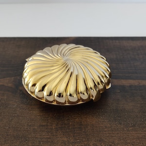 May include: A round, gold-colored compact case featuring a raised, swirling, sunburst-like pattern that radiates from the center to the outer edges. The metallic surface has a polished, reflective finish with deep grooves creating a textured appearance.