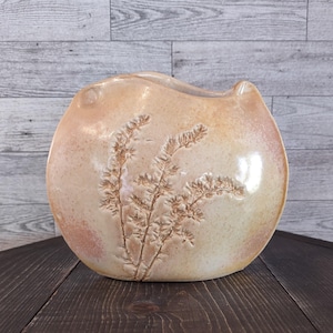 May include: A beige ceramic vase with a rounded shape and a wide opening. The vase features an embossed design of a plant with delicate, feathery leaves. The vase is displayed on a dark wooden surface.