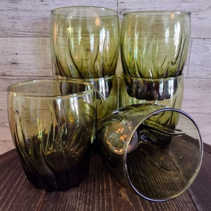May include: A set of five olive green glass tumblers featuring a vertical ribbed texture. The glasses have a rounded shape with a wider top and a slightly tapered base. Four glasses are standing upright, while one glass is lying on its side.