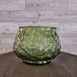 May include: A green glass bowl with a textured, organic pattern. The bowl has a rounded shape and sits on a small base. The glass has a slight transparency, and the bowl is empty.