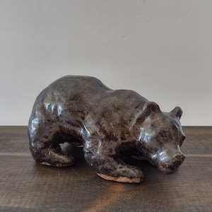 May include: A dark brown ceramic bear figurine in a low, prowling stance. The sculpture features a glossy, uneven glaze with visible texture across the body, rounded ears, and a snout pointed forward, crafted with a solid, heavy appearance.