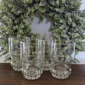 May include: A set of four clear glass tumblers, each featuring a smooth upper cylinder and a textured, ribbed base design. The glasses are arranged in a cluster, showcasing their transparent construction and consistent cylindrical shape.
