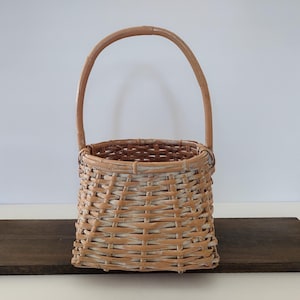 May include: A small, rectangular woven basket with a curved handle. The basket is made of light brown wicker with white accents, and sits on a dark brown surface. The handle is also light brown.